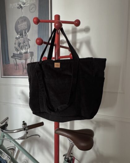 Czarna torba z długimi uchwytami zawieszona na czerwonym stojaku na ubrania, z rowerem w tle.