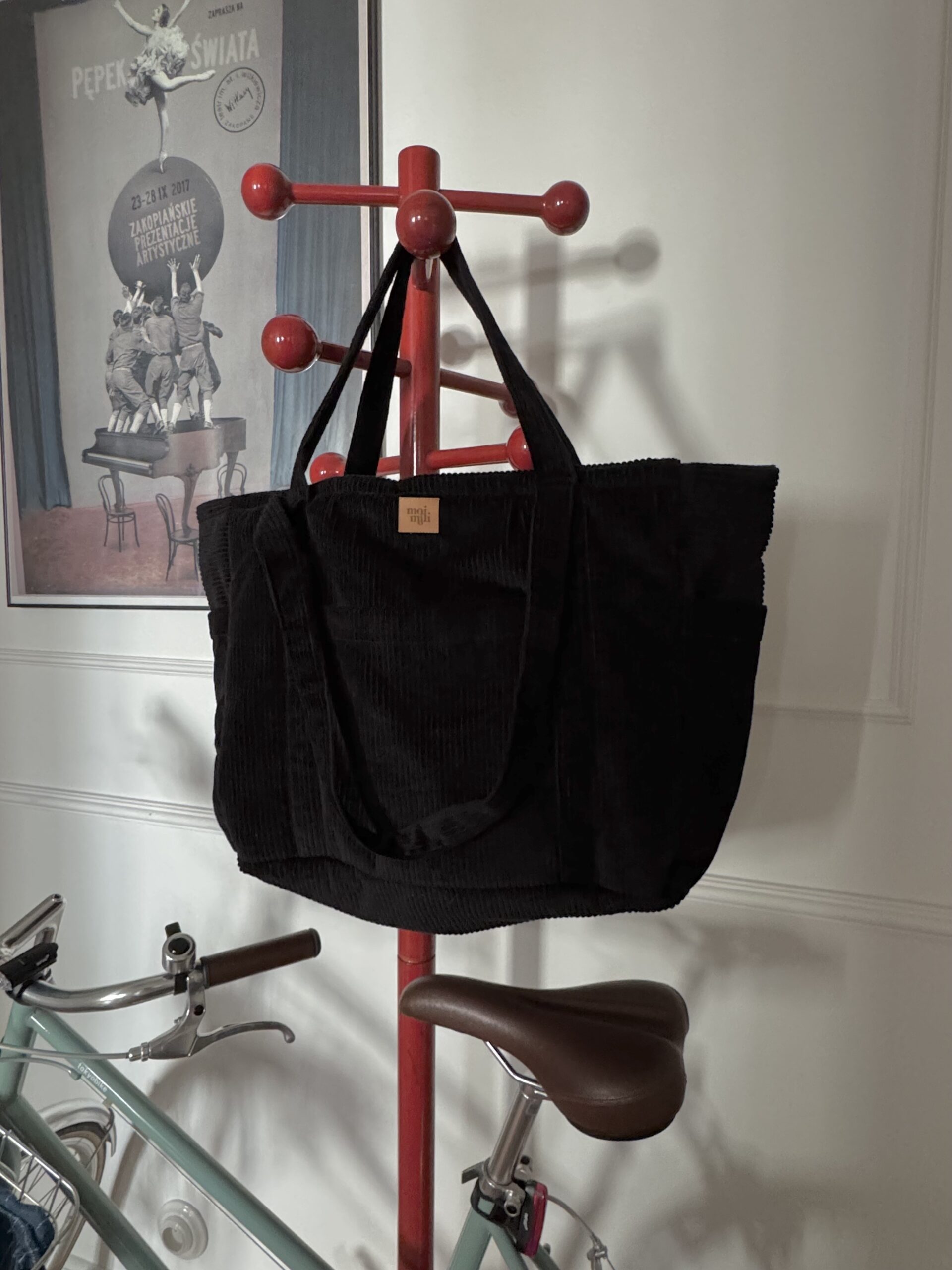 Czarna torba z długimi uchwytami zawieszona na czerwonym stojaku na ubrania, z rowerem w tle.