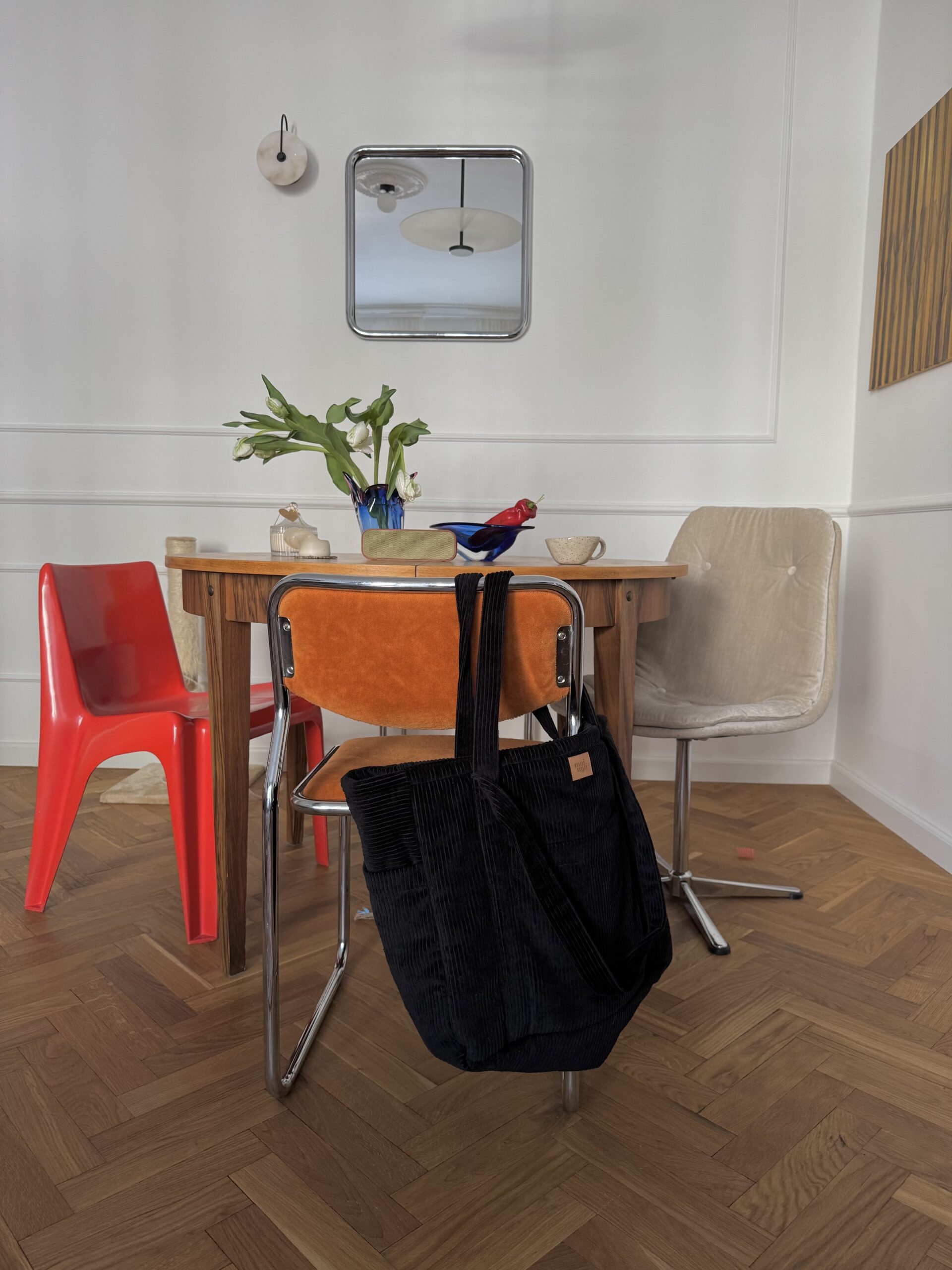 Czarna torba z paskami wisząca na pomarańczowym krześle przy drewnianym stole w jasnym pomieszczeniu z trzema różnymi krzesłami i kwiatami na stole.