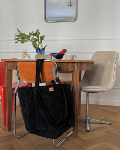 Czarna torba na paskach zawieszona na pomarańczowym krześle przy drewnianym stole w jasnym wnętrzu z lustrzaną lampą i ozdobami na stole.