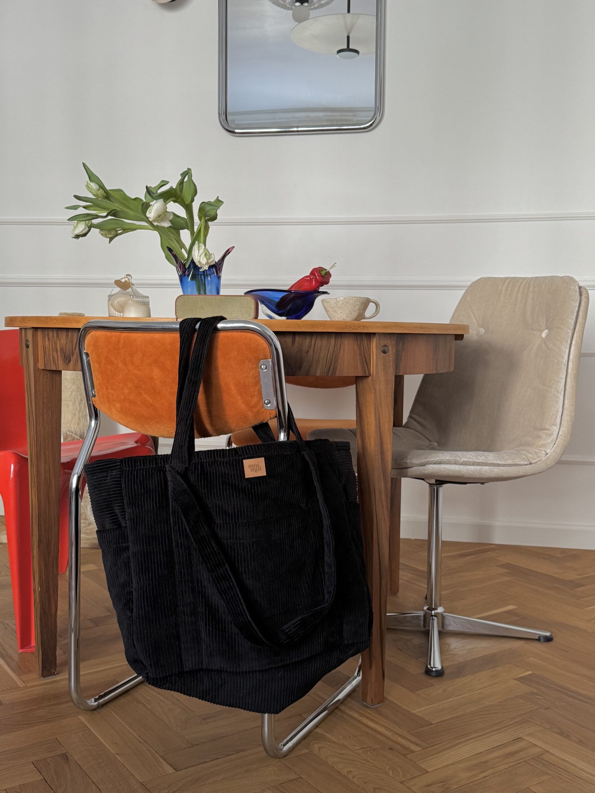 Czarna torba na paskach zawieszona na pomarańczowym krześle przy drewnianym stole w jasnym wnętrzu z lustrzaną lampą i ozdobami na stole.