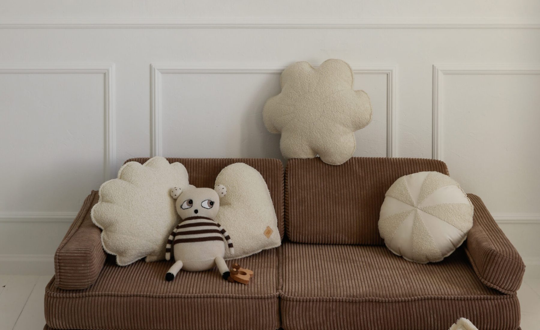 Brązowa kanapa z kremowymi poduszkami i pluszową zabawką na tle białej ściany z panelami